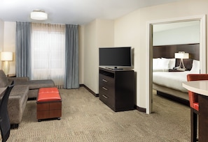 A hotel room with a sofa, a TV on a stand, a desk with a chair, a small table, a mirror, and a bed with a nightstand.