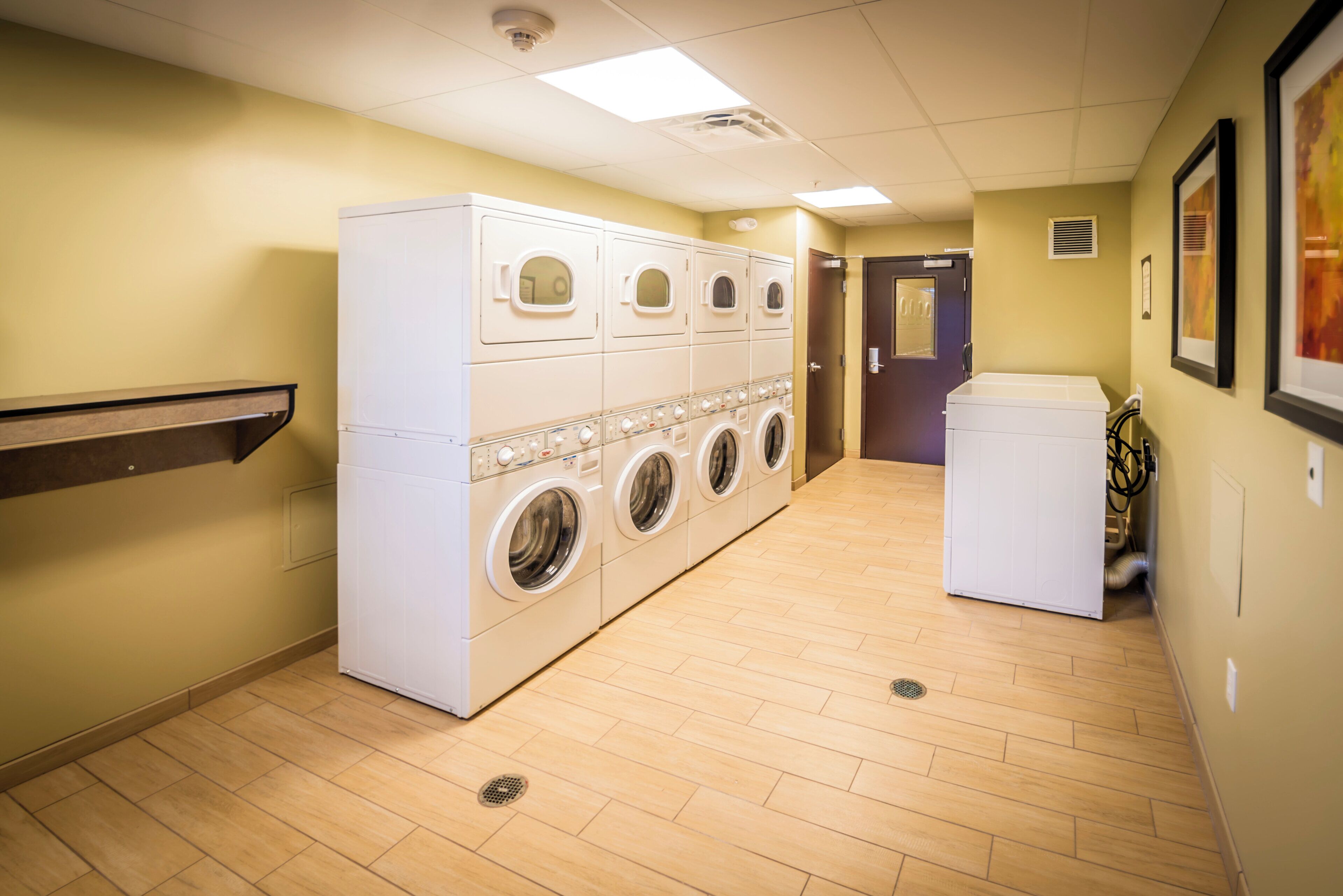 A laundry room with multiple washing machines and dryers, wooden flooring, and a door leading to another room.