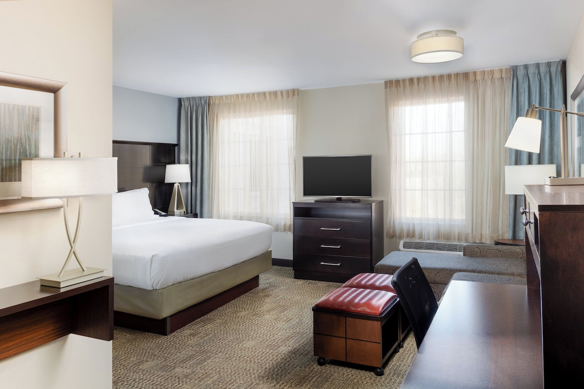 Habitación de hotel con cama, escritorio, televisor, cómoda y ventana con cortinas.