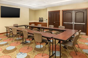 Un salón de conferencias con una larga mesa, sillas, una pantalla de proyector, máquinas de café y una puerta.