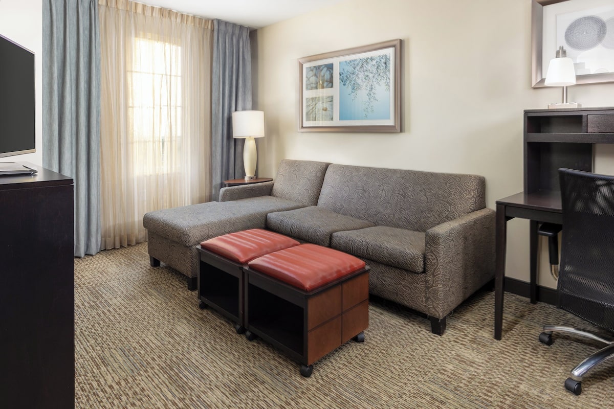 A hotel room with a sofa, ottoman, desk, chair, lamp, and artwork on the wall.