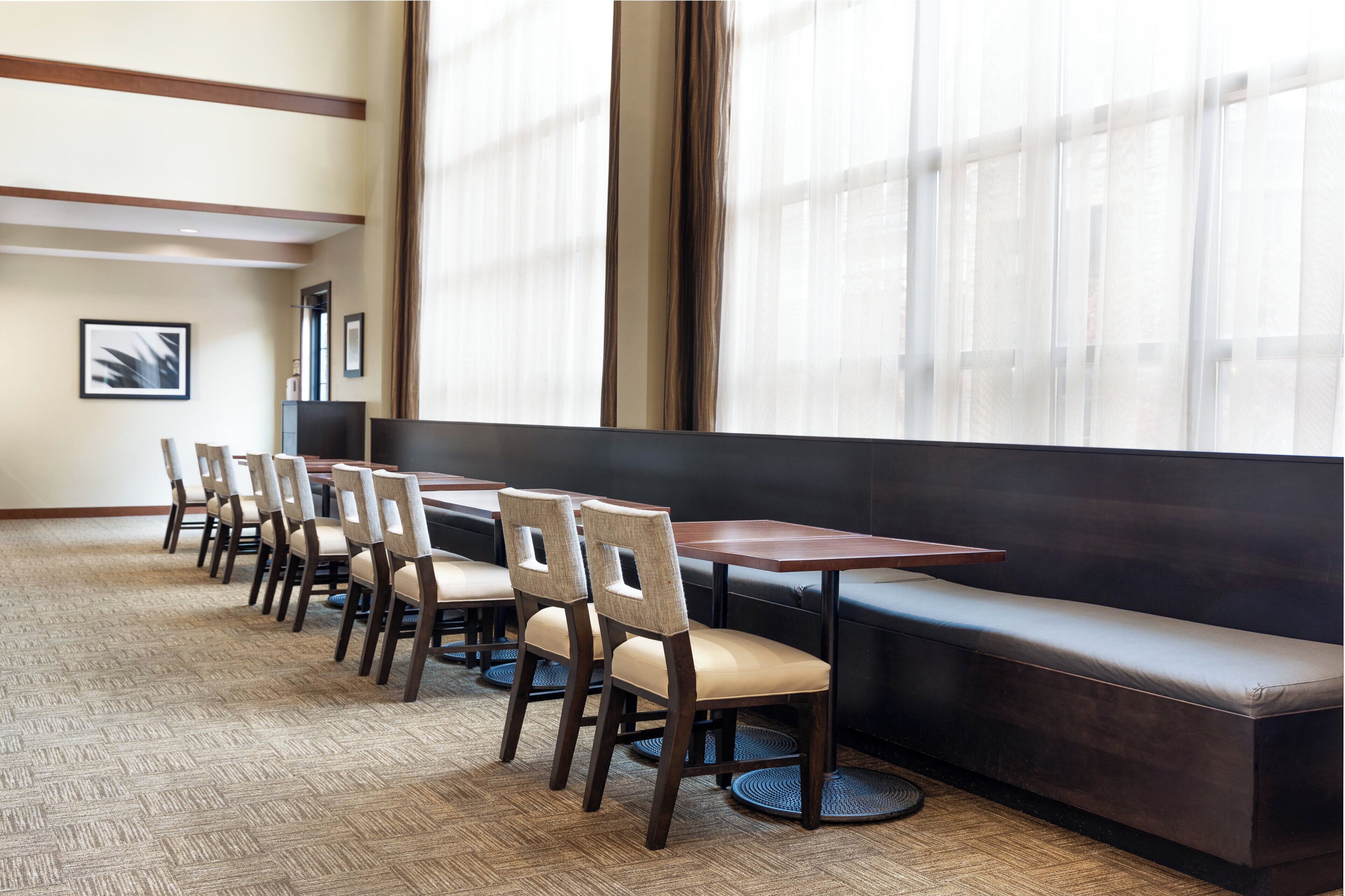A conference room with a long table, several chairs, a bench, and large windows with curtains.