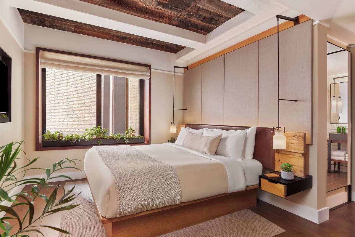 Um quarto moderno com uma cama grande, teto de madeira, janela com plantas e mesa de cabeceira com abajur.