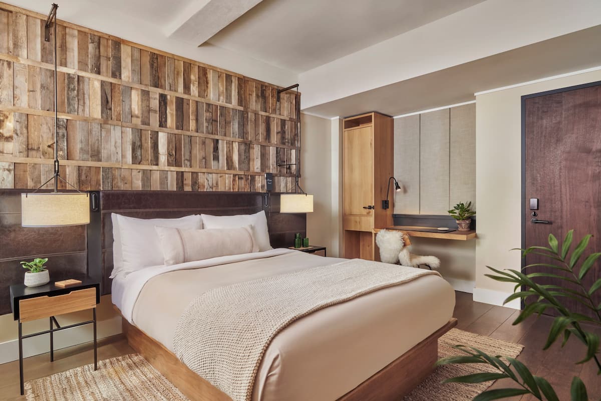 Um quarto com cabeceira de madeira, uma cama com lençóis brancos, uma mesa de cabeceira com abajur, uma pequena mesa com uma planta e um guarda-roupa.