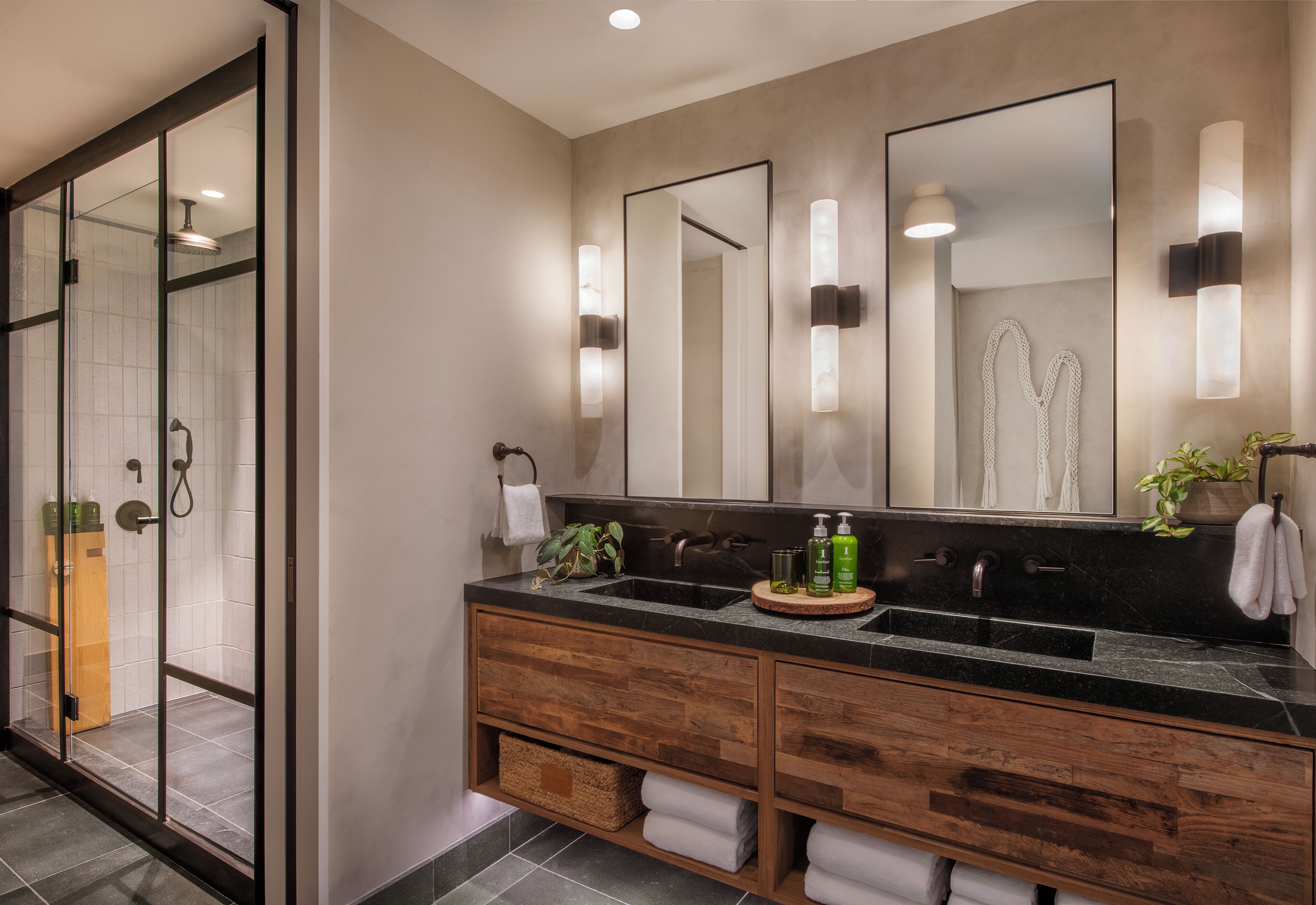 A modern bathroom with a glass-enclosed shower, a wooden vanity with a black countertop, and a large mirror with wall sconces.