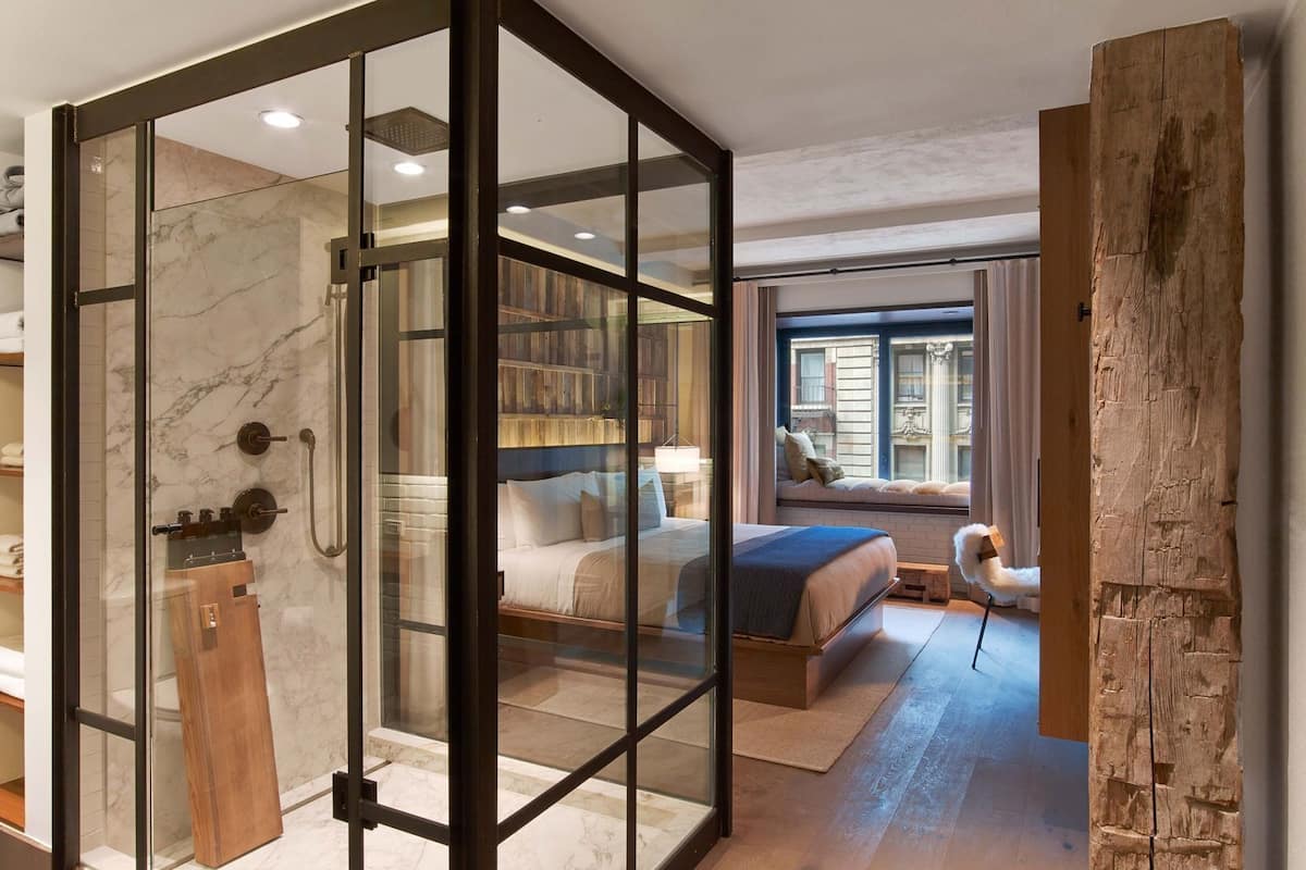 Un dormitorio moderno con separador de vidrio, un banco de madera, cama con vistas y un ventanal grande.
