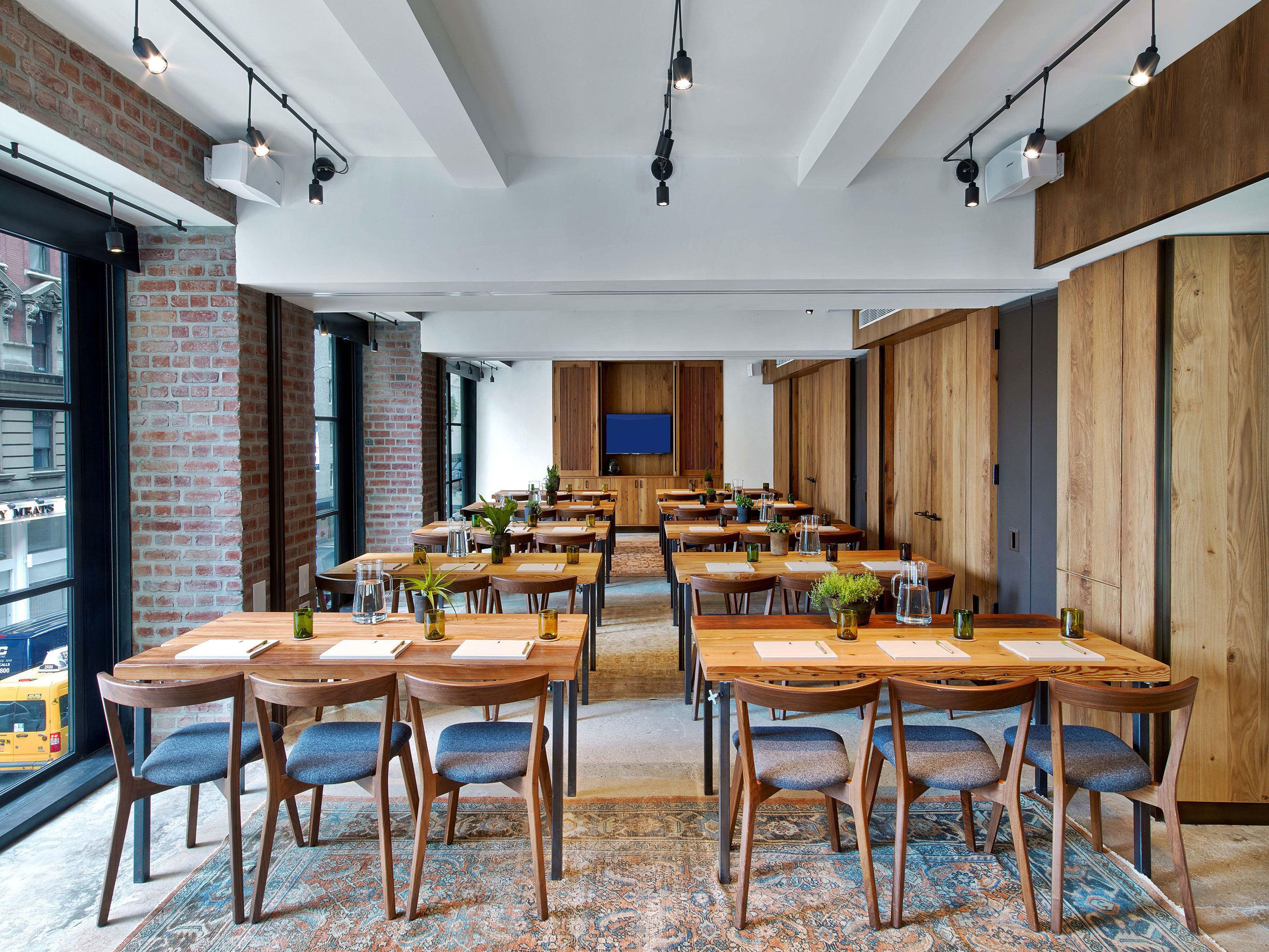 A spacious dining area with multiple wooden tables and chairs, a large TV screen, and a brick wall.