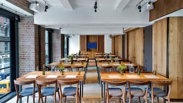 A spacious dining area with multiple wooden tables and chairs, a large TV screen, and a brick wall.