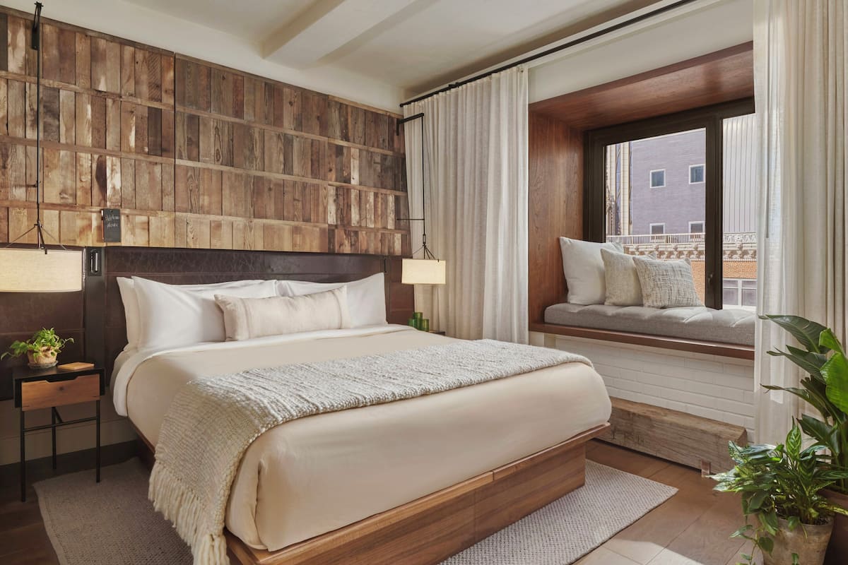 Quarto com uma cama grande, cabeceira de madeira, mesas de cabeceira, um assento junto à janela e vista para edifícios.