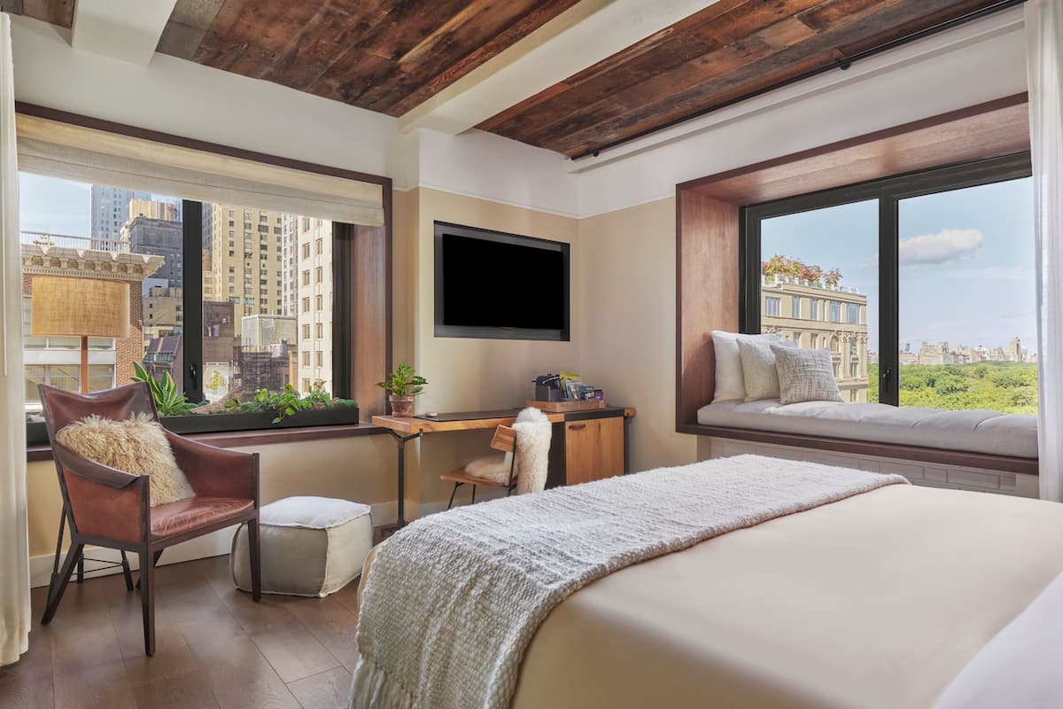 Quarto de hotel com cama, escrivaninha, cadeira, televisão e grandes janelas com vista para a cidade.