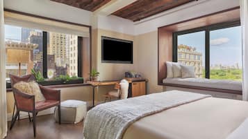 A hotel room with a bed, a desk, a chair, a TV, and large windows with city views.