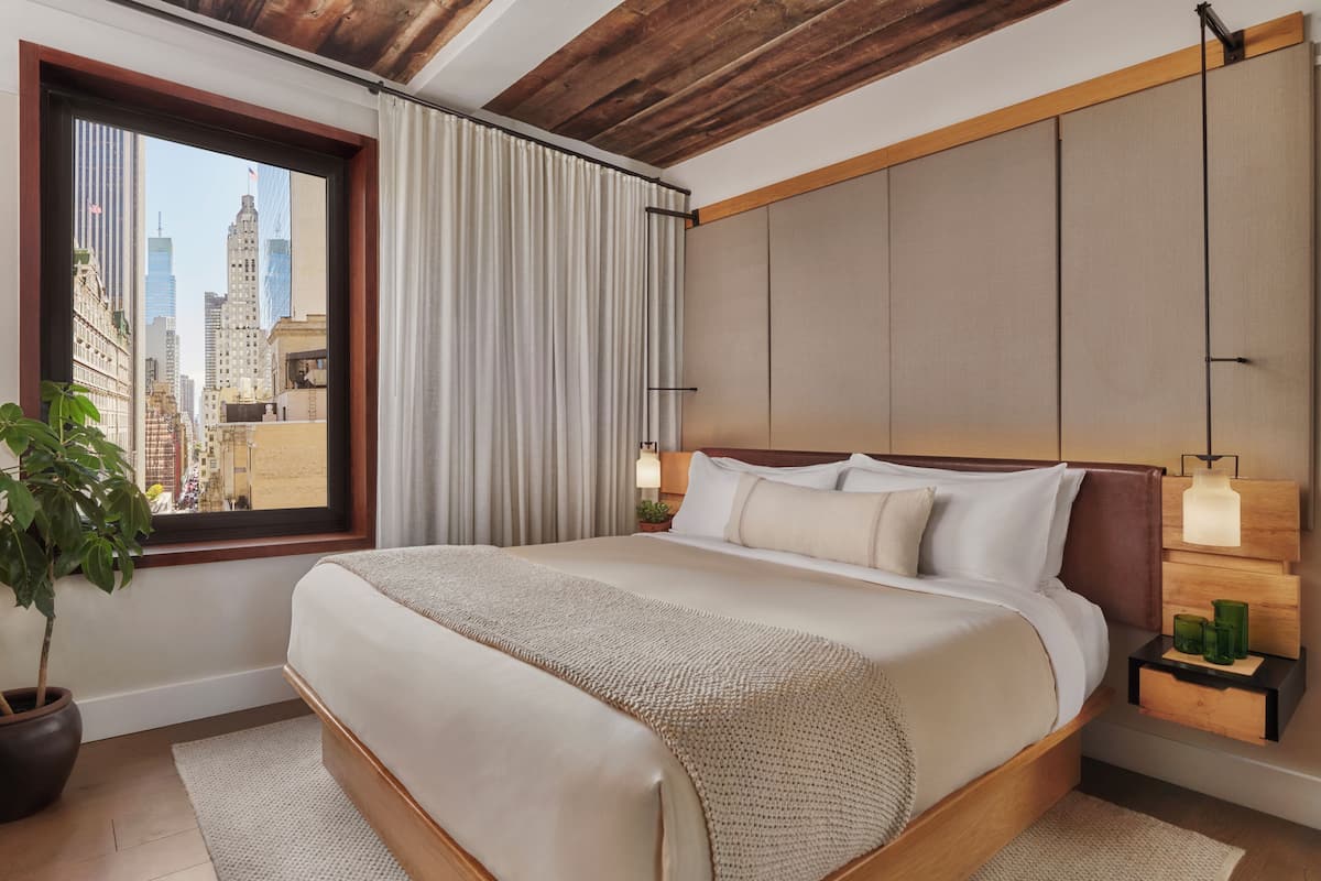 Quarto de hotel com cama grande, janela com cortinas, planta em vaso e vista para a cidade.