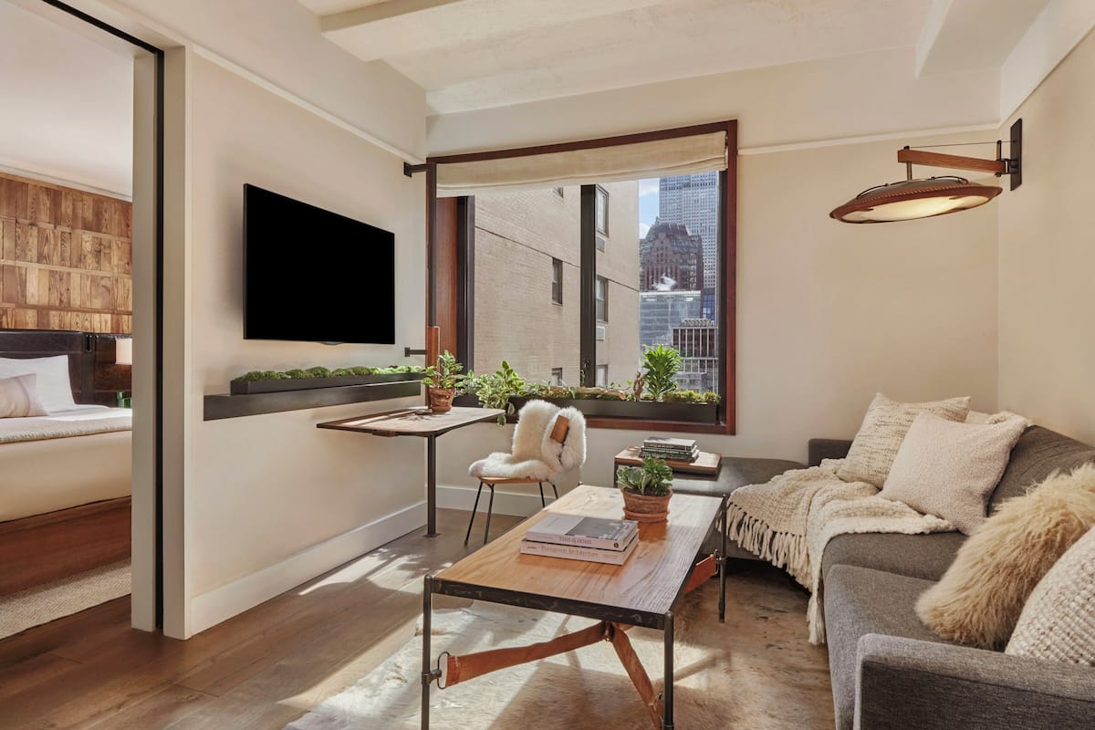 Uma sala de estar moderna com sofá, mesa de centro, TV fixada na parede, janela com vista para edifícios e piso de madeira.