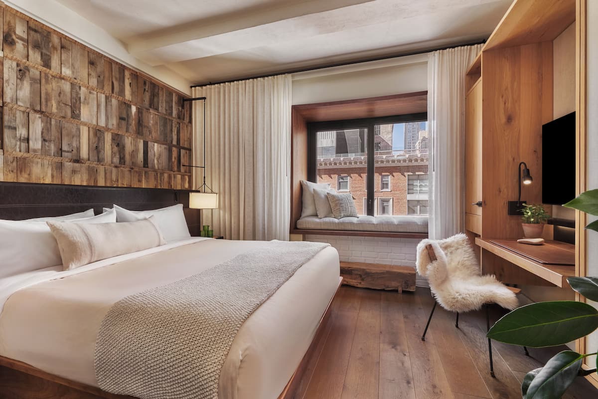 Quarto de hotel com uma cama grande, cabeceira de madeira, assento junto à janela com vista para edifícios, escrivaninha com televisão e uma cadeira.