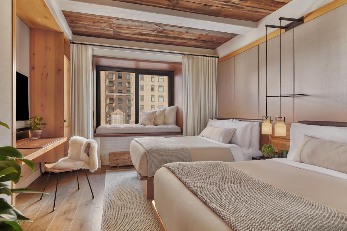 Quarto de hotel com duas camas, teto de madeira, janela com cortinas, televisão e uma pequena mesa com uma planta.