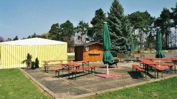 An outdoor seating area with picnic tables and green umbrellas, a small wooden building, and a yellow tent.