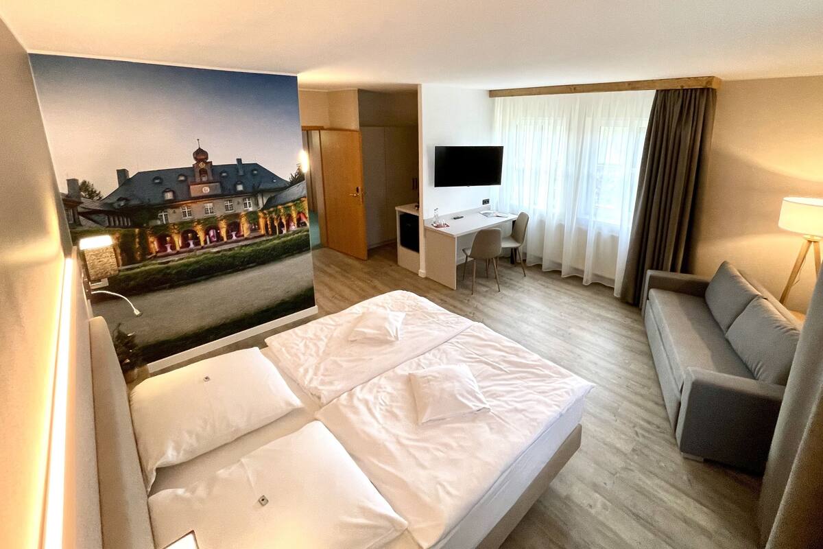A hotel room with a bed, a desk, a chair, a TV, and a view of a building through a window.