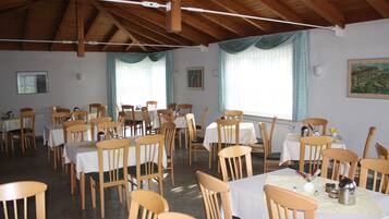 A dining area with wooden tables and chairs, white tablecloths, and a high wooden ceiling.