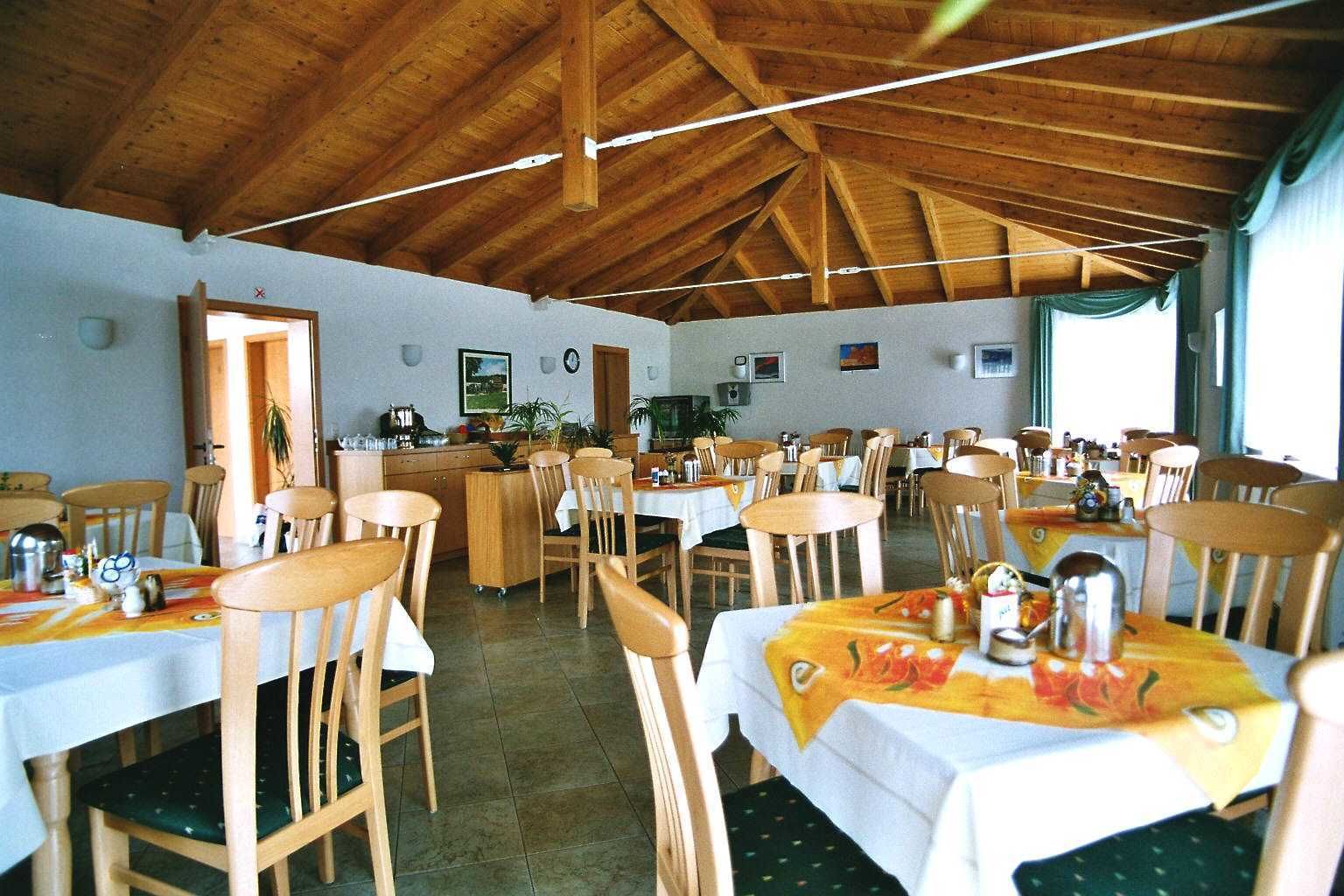 A dining area with wooden tables and chairs, a kitchen in the background, and a high wooden ceiling.