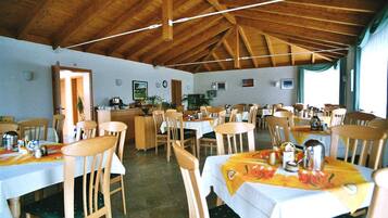 A dining area with wooden tables and chairs, a kitchen in the background, and a high wooden ceiling.