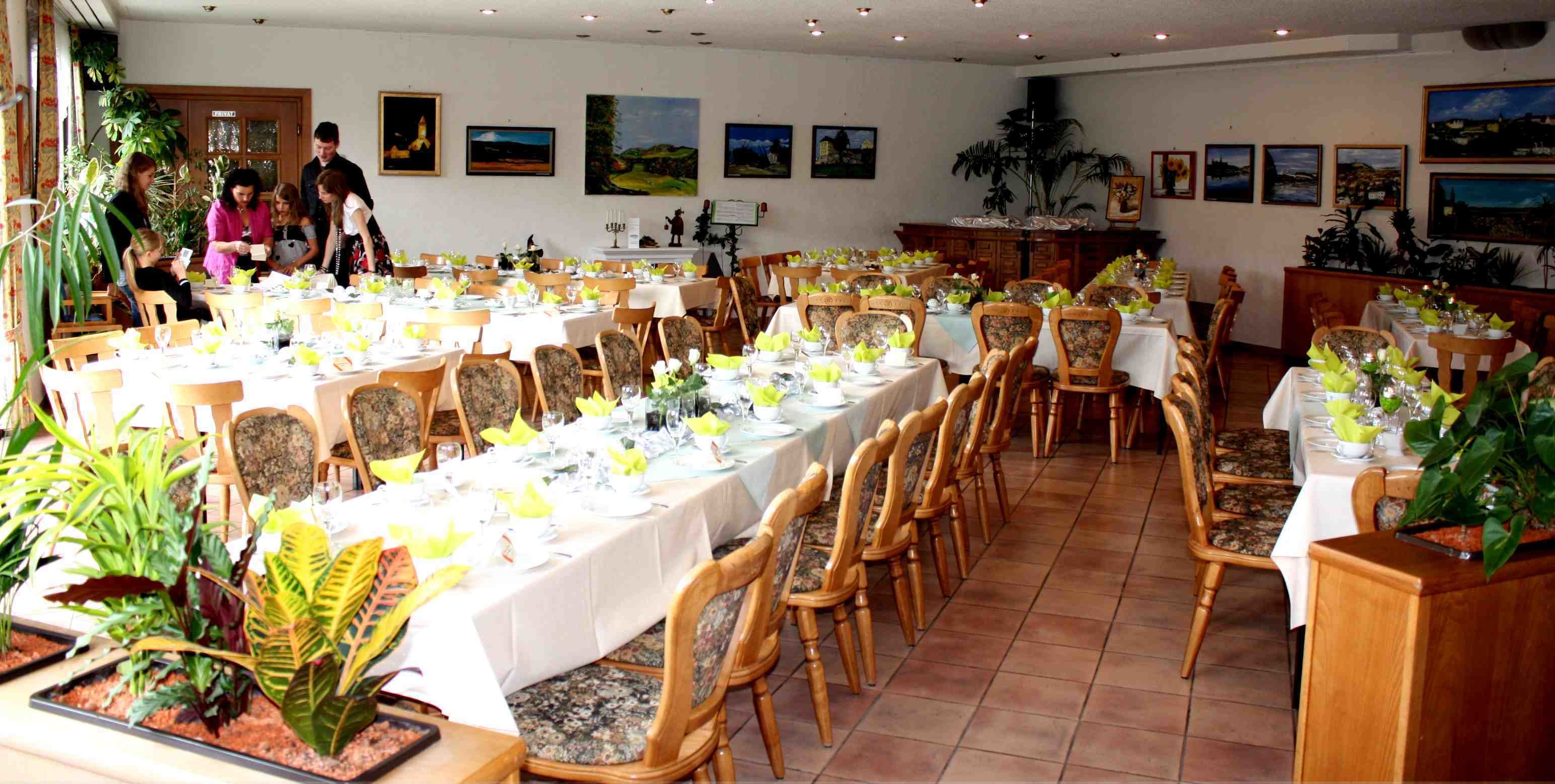 A spacious dining hall with multiple tables set for a meal, surrounded by chairs and potted plants.