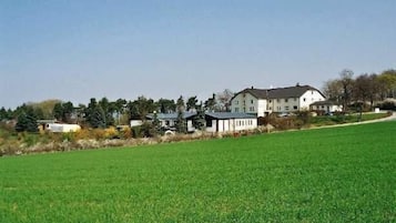 A large white building with multiple windows and a flat roof, surrounded by a green field and other buildings in the background.