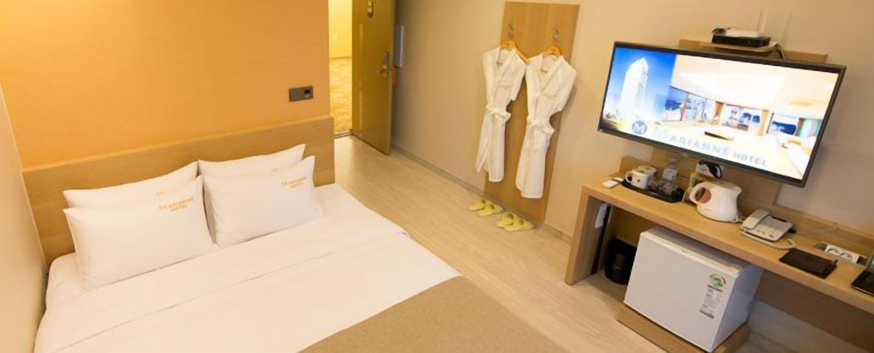 A hotel room with a bed, a television, a small refrigerator, and a wooden side table with a phone and a kettle.