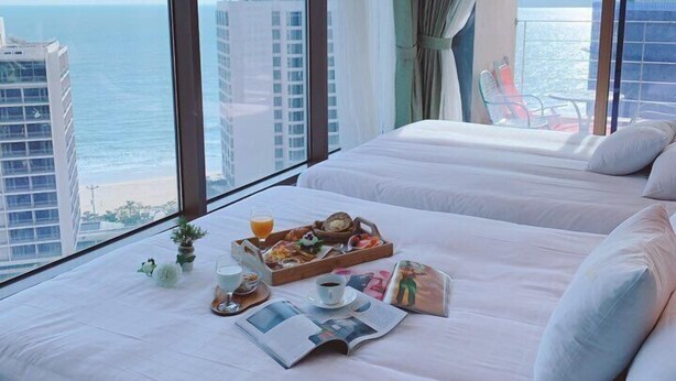 Habitación de hotel con dos camas, una bandeja de desayuno, una revista y vistas al mar y a edificios.