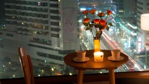 Habitación de hotel con una mesa dispuesta para dos, un jarrón con flores y vistas a un paisaje urbano nocturno.