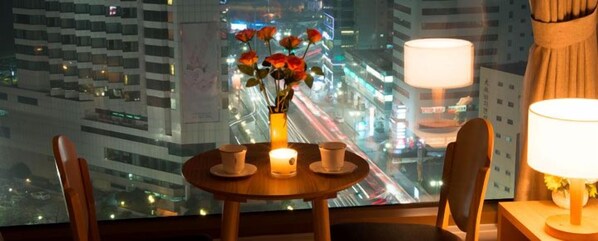 Habitación de hotel con una mesa dispuesta para dos, un jarrón con flores y vistas a un paisaje urbano nocturno.