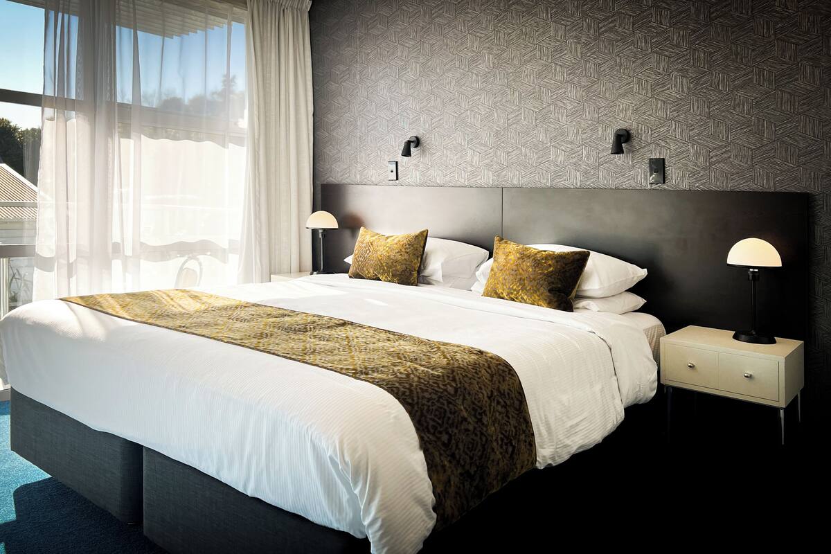 Quarto de hotel moderno com uma cama grande, duas mesas de cabeceira com luminárias e uma parede estampada.