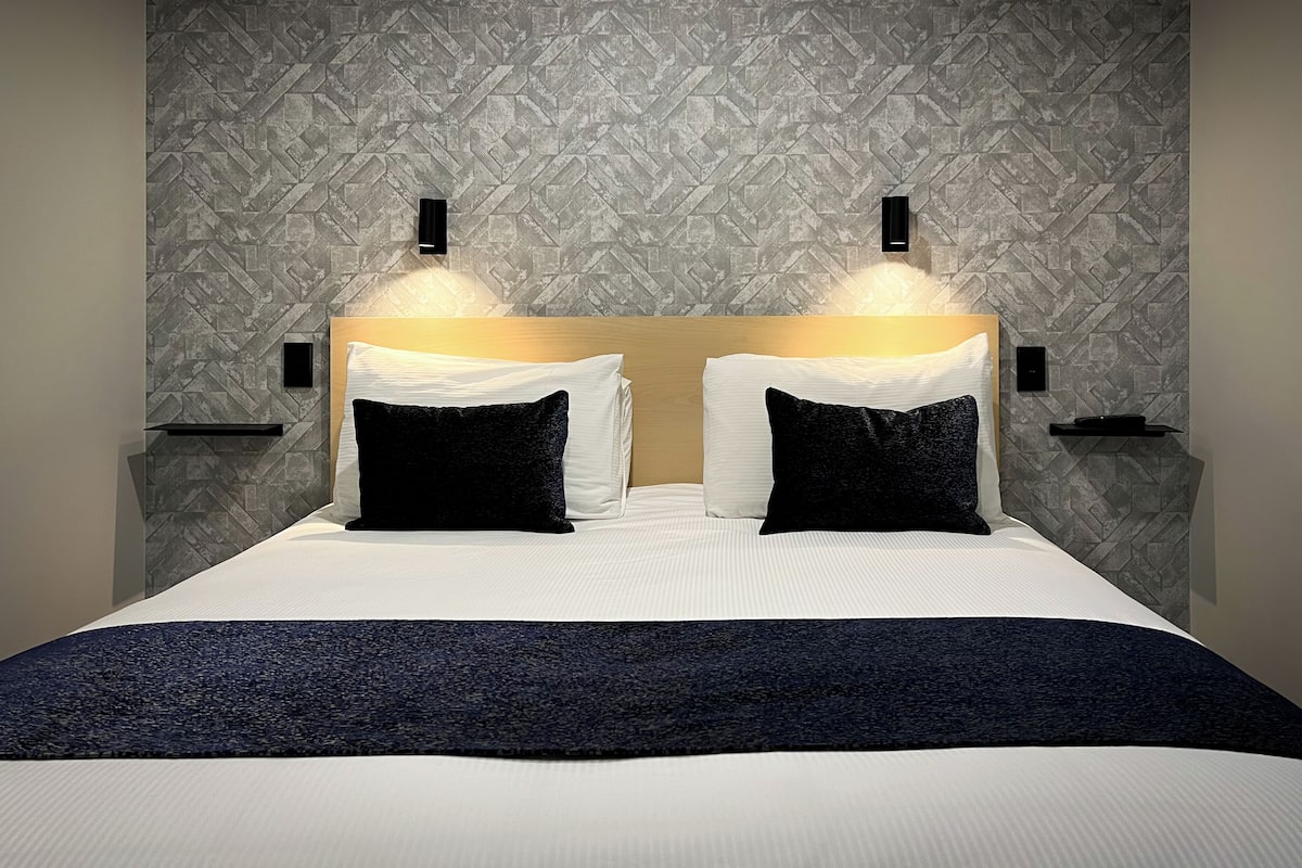 Quarto de hotel moderno com uma cama grande, duas mesas de cabeceira e uma parede com textura.