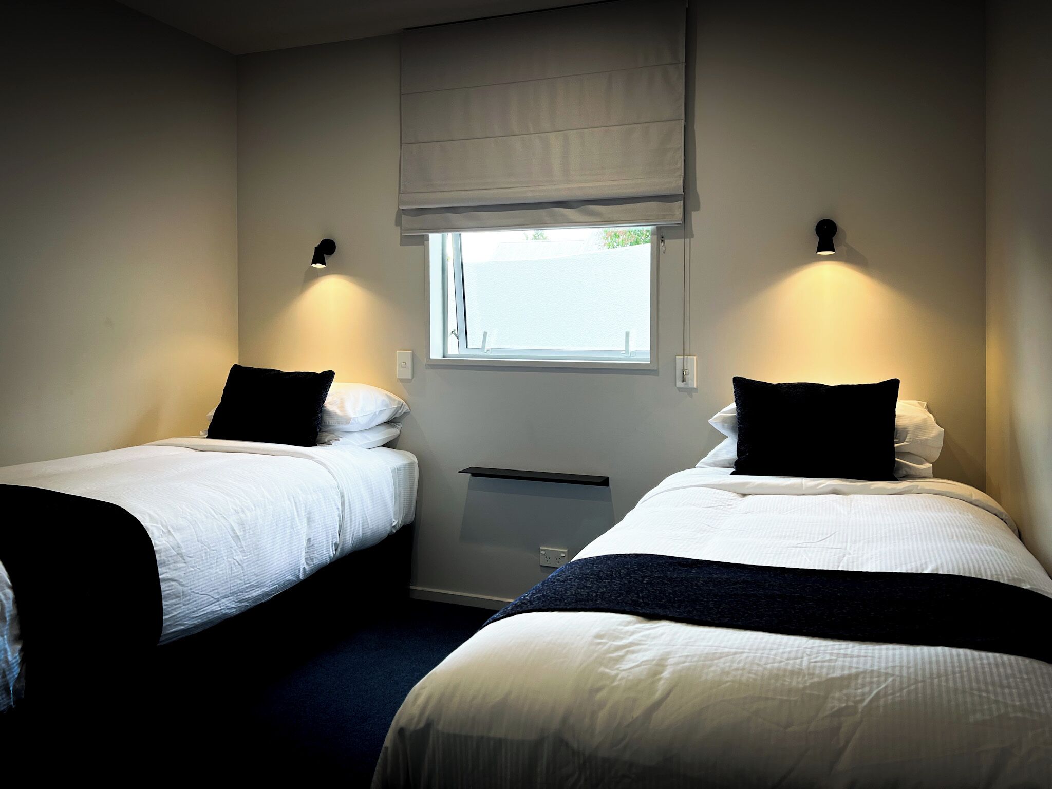 Ein Schlafzimmer mit zwei Betten, jeweils mit weißer Bettwäsche und schwarzen Kissen, einem Fenster mit Jalousie und Wandleuchten.