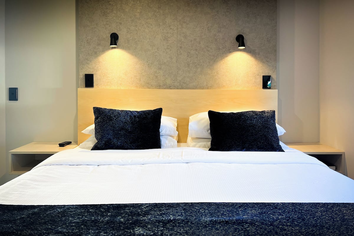 Quarto de hotel com uma cama grande, duas almofadas pretas, uma cabeceira de madeira e duas luzes fixadas na parede.