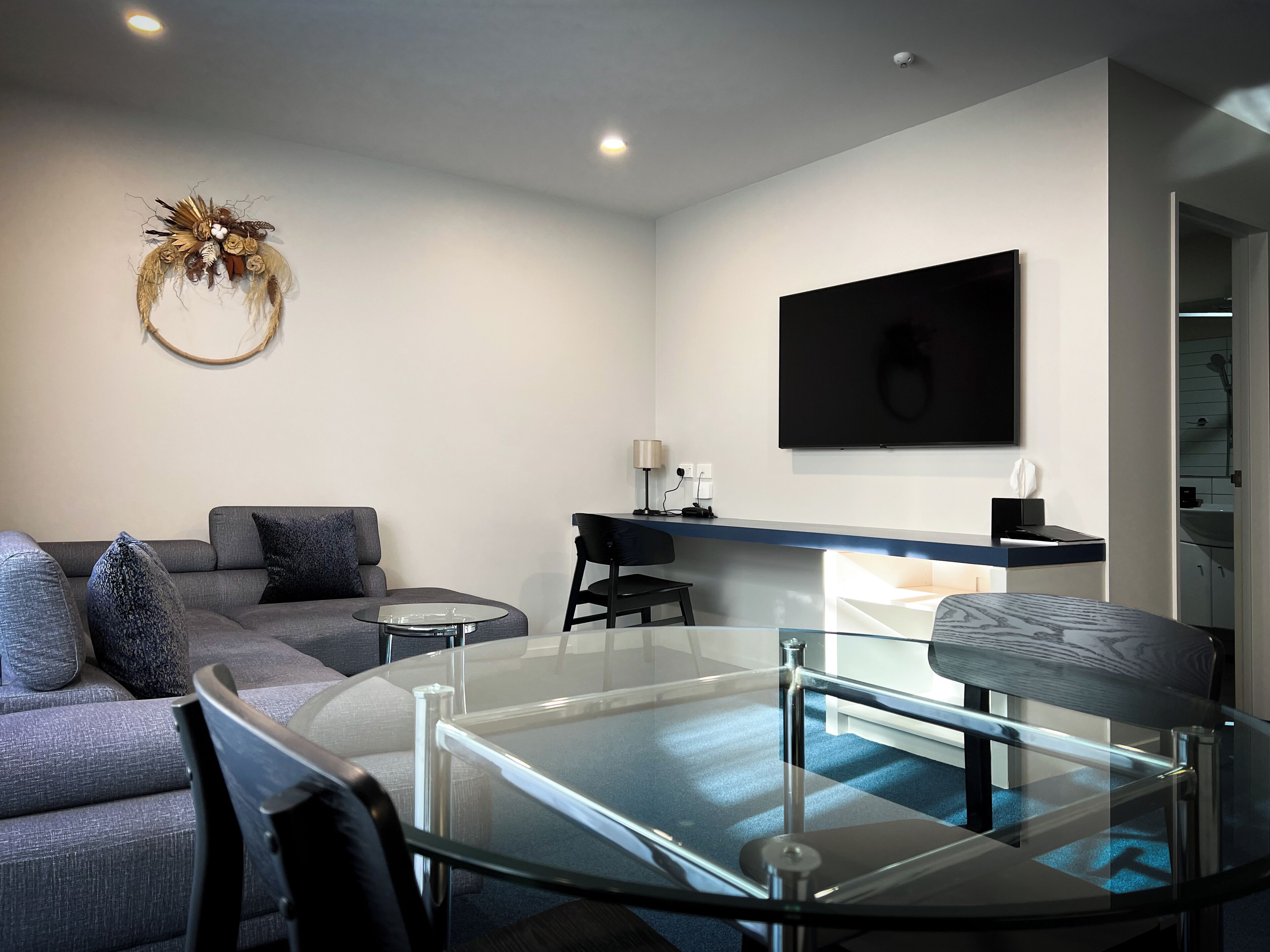 A modern living room with a glass coffee table, grey sofa, and a decorative wall piece.