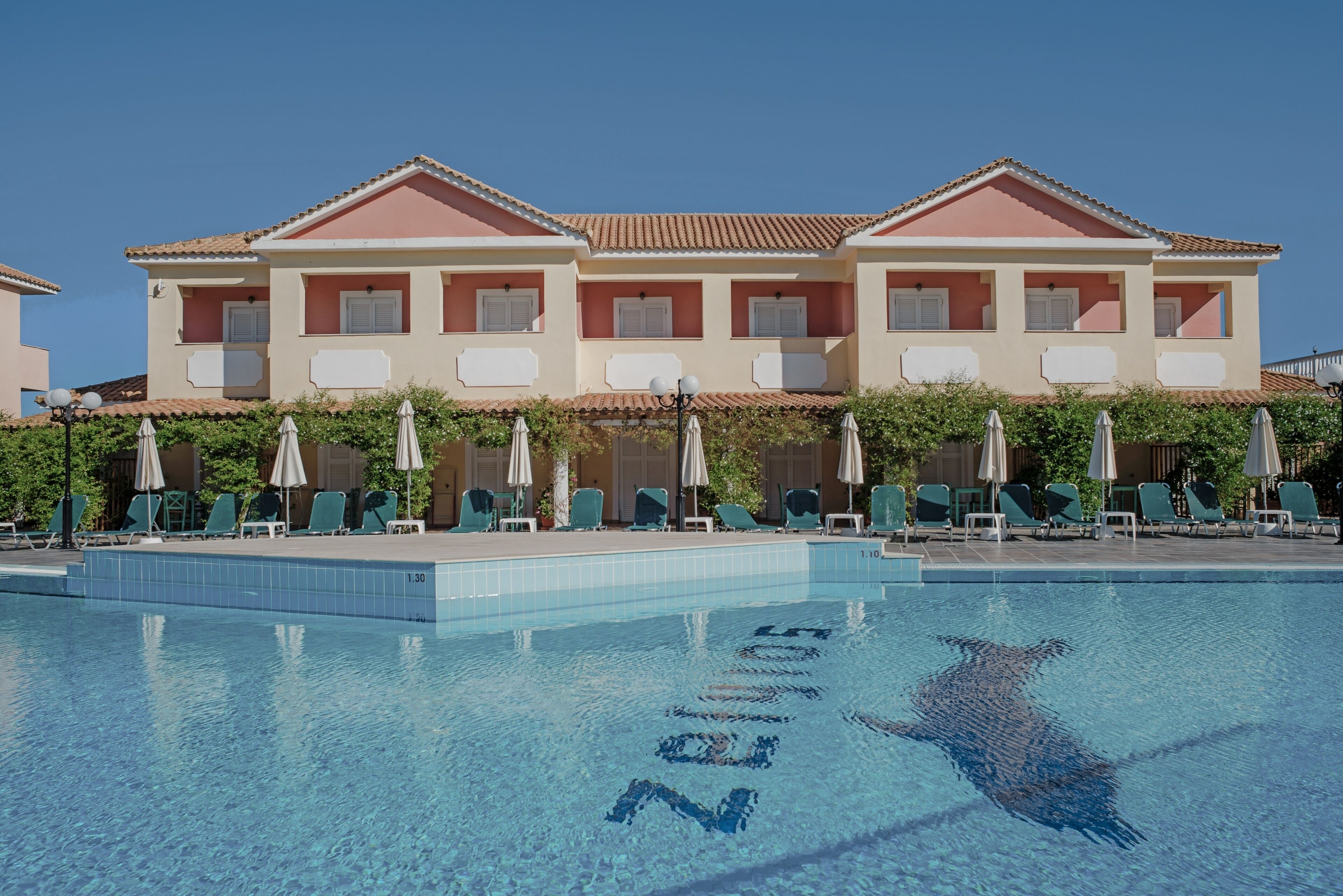 Una piscina con lettini e ombrelloni, un edificio con tetto in tegole e un cielo azzurro limpido.