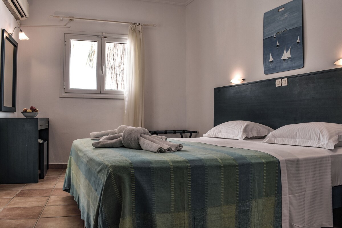 A bedroom with a bed, a nightstand, a window with curtains, a wall-mounted artwork, and a wall-mounted light fixture.