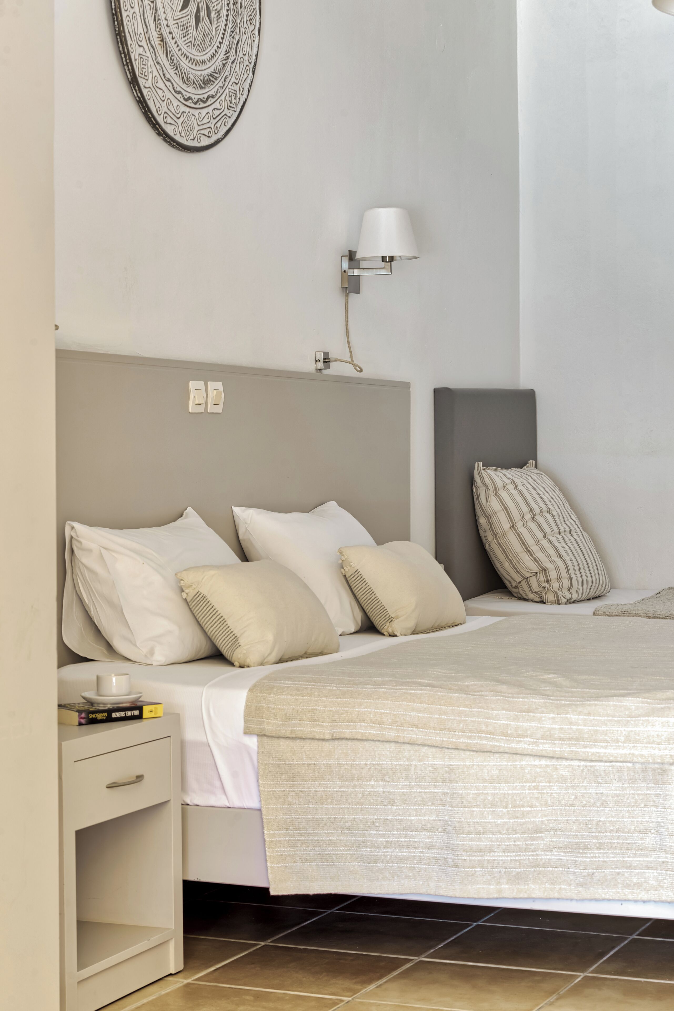 Una cama bien hecha con cabecera gris, una lámpara de noche y una mesita de noche con un libro y una taza.