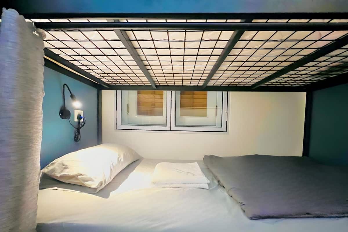 Una habitación pequeña con una cama individual y una litera, una ventana con persianas, una lámpara en la pared y un enchufe.