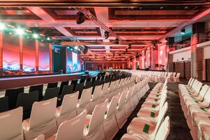 A conference room with rows of white chairs, a stage with a large screen, and a ceiling with lighting fixtures.