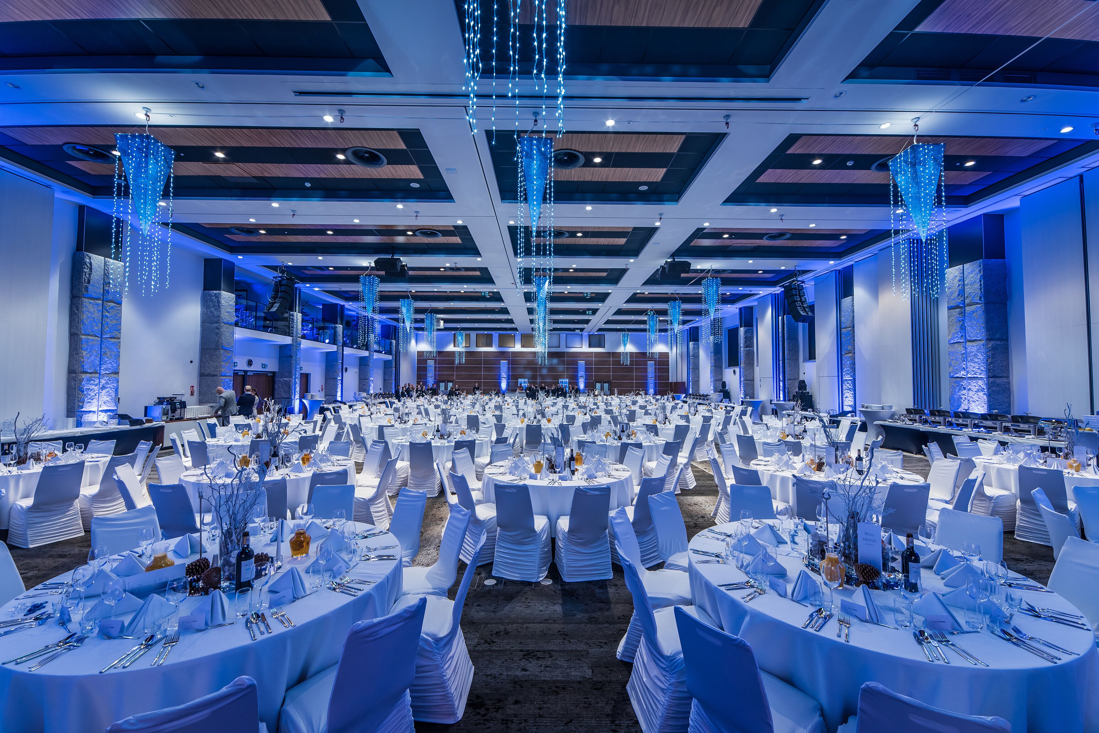 Un gran salón de banquetes con mesas redondas dispuestas para comer, sillas blancas e iluminación azul.