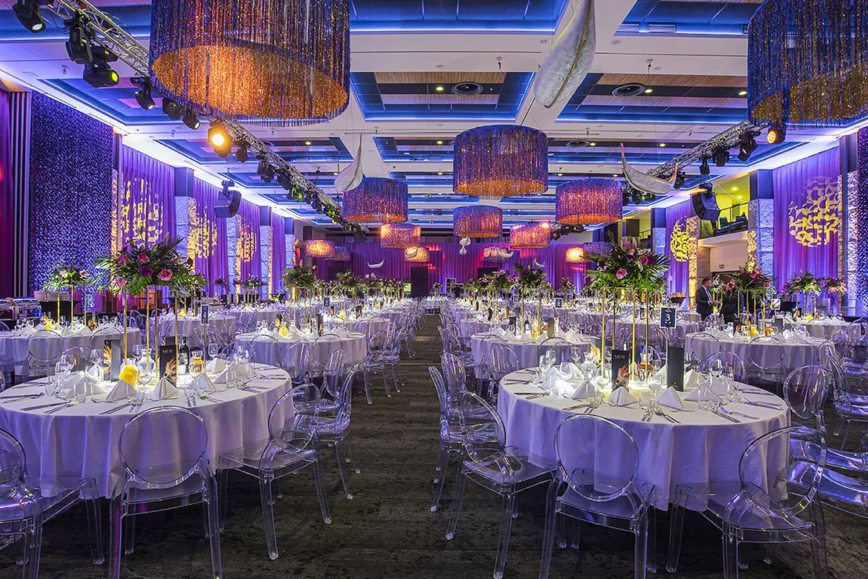 Une salle de banquet avec des tables rondes, des chaises transparentes et des rideaux violets.