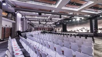 A conference room with rows of white chairs, a stage with a backdrop, and modern ceiling lighting.