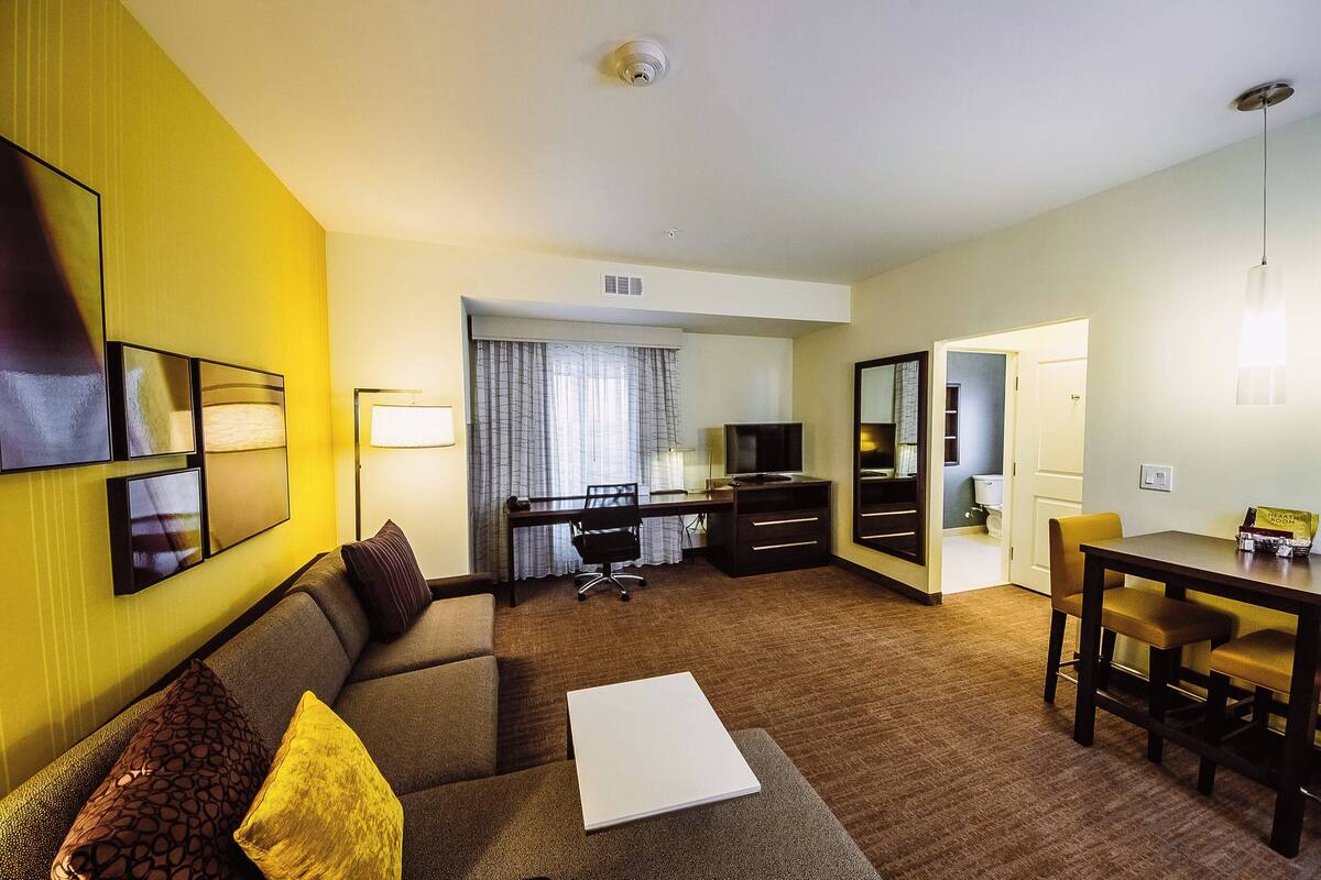 A hotel room with a sofa, a dining table with chairs, a desk with a chair, a television, and a mirror.
