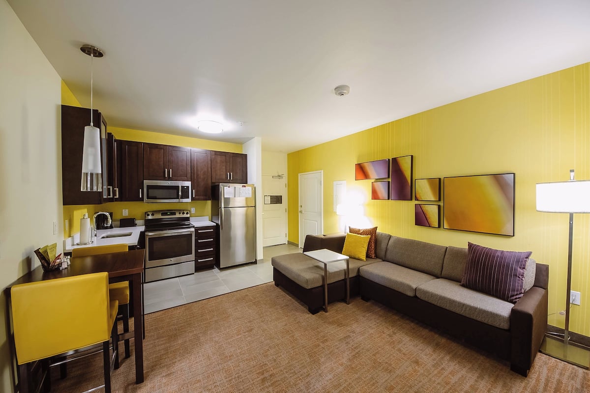 A modern kitchen with stainless steel appliances, a dining area with a yellow chair, and a living room with a sofa and abstract wall art.