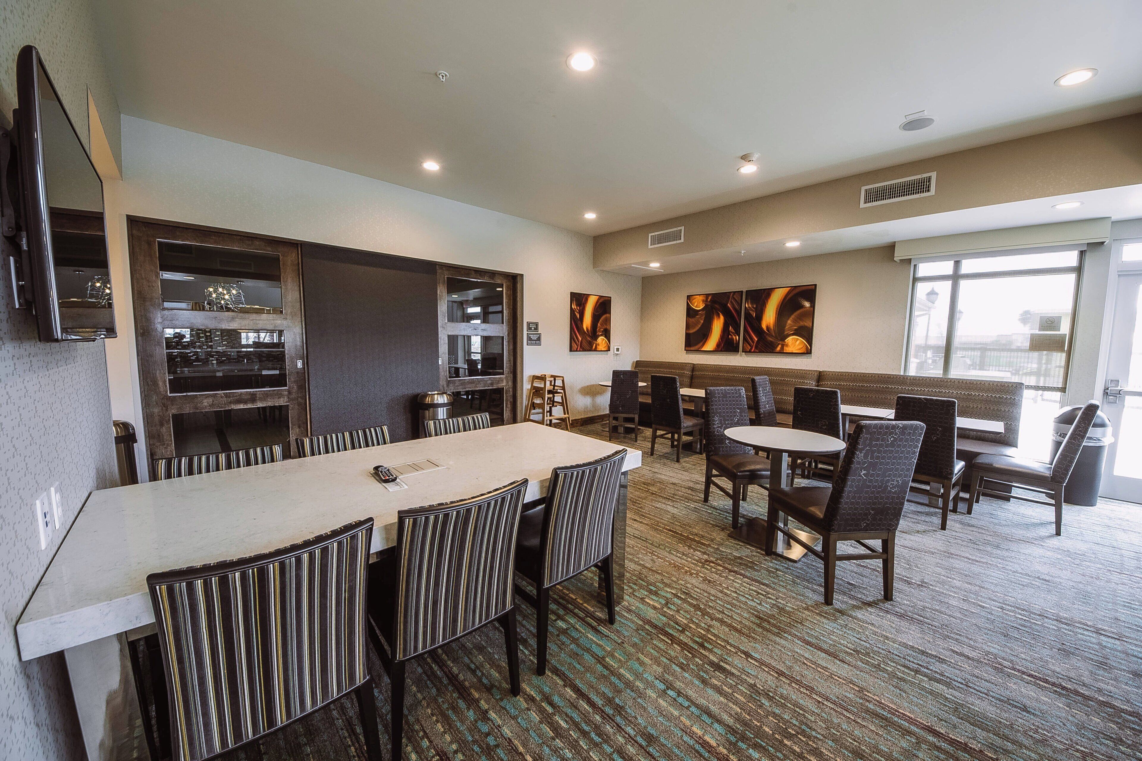 Comedor de hotel con una larga mesa común, varias mesas más pequeñas y diversas opciones de asiento, incluyendo cabinas y sillas.