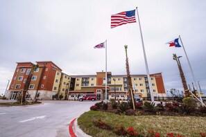 Un hôtel à plusieurs étages avec une entrée principale, des drapeaux américains et du Texas, et un espace de stationnement bordé d’une bordure rouge.