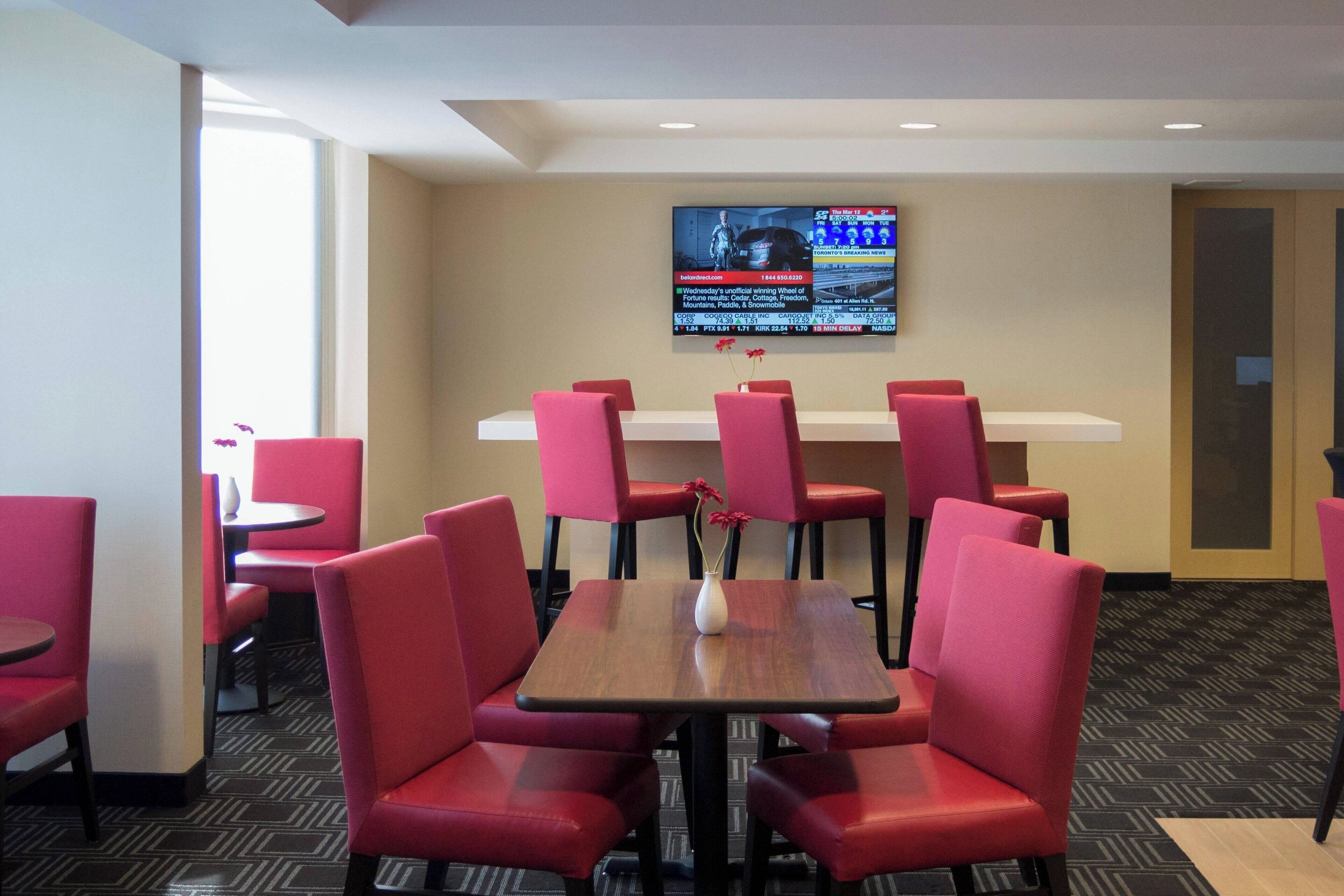 A hotel room with a large flat-screen TV mounted on the wall, a wooden table with a vase of flowers, and red upholstered chairs.