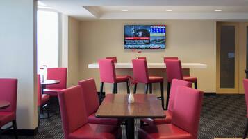A hotel room with a large flat-screen TV mounted on the wall, a wooden table with a vase of flowers, and red upholstered chairs.