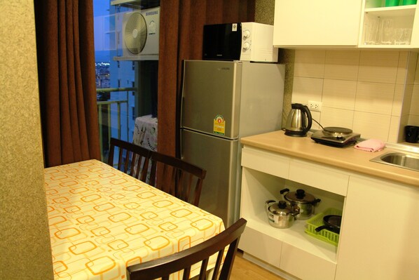 A compact kitchen with a refrigerator, microwave, and a small countertop. There is a dining table with a patterned tablecloth and a chair. - Lumpini Ville Naklua Good Luck (Pattaya)